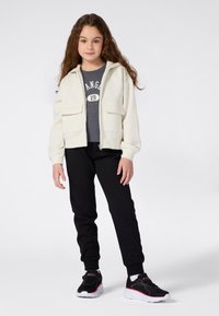 Een jong meisje staat in een crèmekleurige ritsjack, een donkergrijs graphic T-shirt, zwarte joggers en zwarte sneakers met roze accenten.