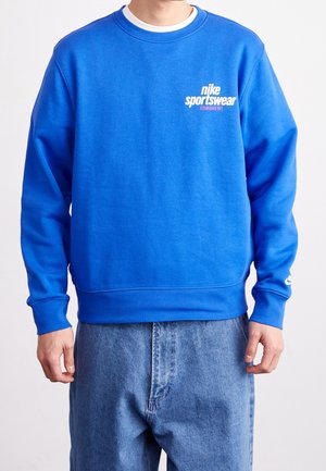 Mężczyzna noszący jaskrawoniebieską bluzę Nike Sportswear z białym napisem oraz jasnoniebieskie jeansy, stojący na jednolitym tle.