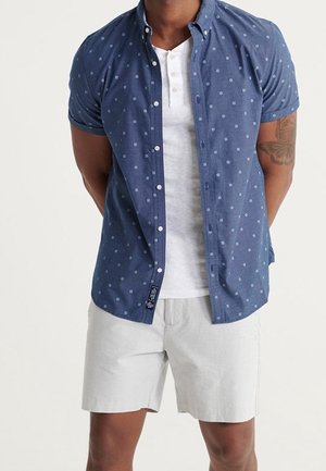 Mężczyzna ubrany w biały, zapinany sweter henley pod niebieską koszulą na krótki rękaw z małym wzorem w kwadraty oraz jasne spodenki.