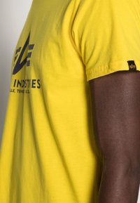 Žluté tričko s krátkým rukávem vyrobené z bavlny. Obsahuje grafické logo v tmavě modré barvě a malou černou cedulku na rukávu. Hladká textura.