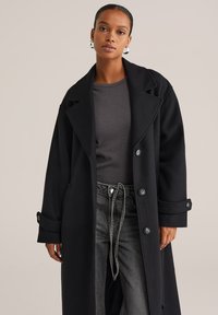 Cappotto di lana nera con grandi rever, chiusura a bottoni e polsini arrotolati, indossato sopra un top grigio e abbinato a jeans scuri.