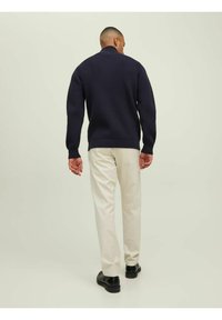 Suéter de punto navy con cuello alto, combinado con pantalones beige claros y zapatos negros, visto desde la espalda contra un fondo liso.