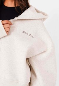 Ljusgrå hoodiet med dragkedja fram, mjuk fleece-textur, ribbade muddarna och broderad "Sixth June"-logotyp på bröstet.