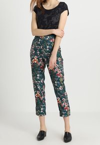 Camiseta negra con un patrón floral, combinada con unos pantalones verdes de longitud tobillo que presentan un vibrante diseño floral. La modelo lleva puestas unas zapatillas negras sin cordones.