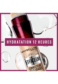 Le tube de correcteur Maybelline présente un corps transparent, un capuchon applicateur en brosse et des accents métalliques. L'étiquette comprend "Instant Anti-Age" et "Hydratation 12 Heures."