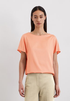 Short-sleeve coral top with a wide boat neckline, soft fabric, flowy shape, paired with light beige high-waisted trousers.