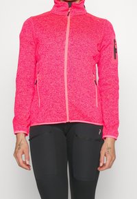 Kobieta ubrana w jasnoróżową polarową kurtkę na zamek błyskawiczny z dwoma bocznymi kieszeniami oraz czarne legginsy z kieszeniami na udach z zamkiem.