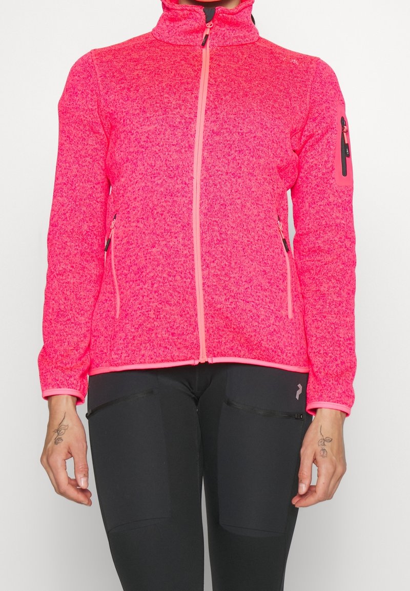 Kobieta ubrana w jasnoróżową polarową kurtkę na zamek błyskawiczny z dwoma bocznymi kieszeniami oraz czarne legginsy z kieszeniami na udach z zamkiem.