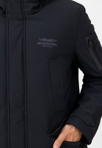 Schwarze Bomberjacke mit glatter Textur, ausgestattet mit einer Reißverschlusstasche am Ärmel und Logodetails auf der Brust. Mehrfach Taschen-Design.