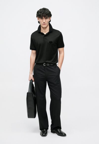 Juoda polo marškinėliai su trumpu apykakle ir siuvinėta logotipu, derinami su juodomis kelnėmis ir elegantiška juoda rankine, fone baltas paviršius.