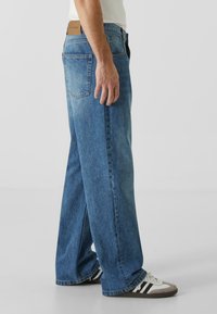 Široke jeans hlače iz modrega denima s pošvikanim videzom, sprednjima in zadnjima žepoma ter usnjenim oznako na pasu. Ujemajo se s črnimi in belimi supergami.
