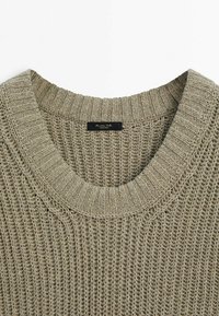Lys taupe strikket sweater med bred rund halsudskæring. Funktioner et struktureret stingmønster og et mærke syet ved halsen. Blød, afslappet design.