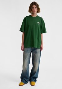 Green oversized t-shirt with a round neck and short sleeves, paired with wide-leg faded blue jeans and yellow sneakers.
