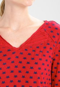 Top in tessuto rosso con scollo a V, dettagli in pizzo intricato al collo e un motivo di piccole silhouette di gatti blu navy.