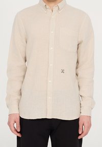 Uomo che indossa una camicia beige con bottoni, a maniche lunghe, dotata di taschino e di un piccolo testo ricamato in nero vicino alla parte inferiore frontale.
