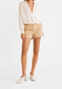 Pantalones cortos beige combinados con una camisa blanca de manga larga. La camisa tiene un escote en V y un ajuste holgado. Los zapatos son de color blanco roto con un tacón bajo.