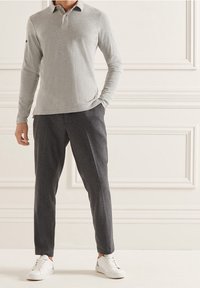 Mann trägt ein hellgraues Langarm-Polo, dunkelgraue maßgeschneiderte Hosen und weiße Sneaker und steht vor einer weißen Verkleidungswand.