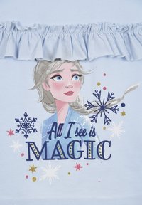 Camiseta azul cielo con cuello con volantes que presenta una impresión de la cara de un personaje, el texto "Todo lo que veo es MAGIA" y motivos de copos de nieve.