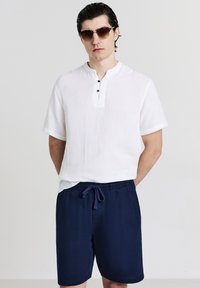 Uomo che indossa occhiali da sole tartarugati, una camicia bianca a maniche corte con bottoni e pantaloni corti blu navy con cordino, in piedi davanti a uno sfondo neutro.