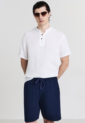 Homme portant des lunettes de soleil en écaille de tortue, une chemise blanche à manches courtes boutonnée et un short bleu marine avec cordon de serrage, se tenant devant un fond uni.