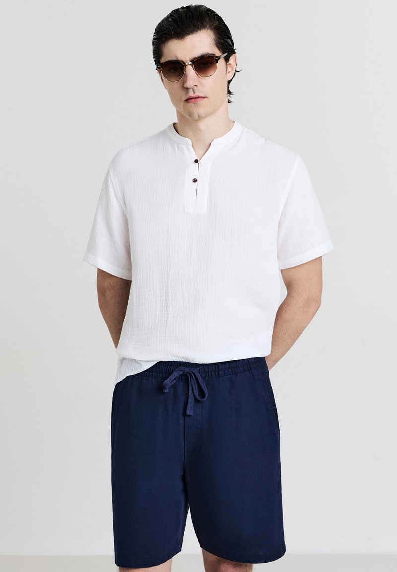 Uomo che indossa occhiali da sole tartarugati, una camicia bianca a maniche corte con bottoni e pantaloni corti blu navy con cordino, in piedi davanti a uno sfondo neutro.