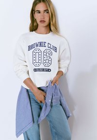 Sudadera blanca con la inscripción "Brownie Club 06 College Dept." en color marino y estrellas. El modelo la combina con vaqueros azul claro y sostiene una camisa a rayas.