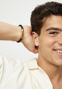 Pulsera de cuero trenzado negro con un cierre dorado. El modelo lleva una camisa de color claro, sonríe, tiene el cabello corto y oscuro y adopta una postura relajada.