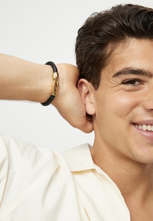 Pulsera de cuero trenzado negro con un cierre dorado. El modelo lleva una camisa de color claro, sonríe, tiene el cabello corto y oscuro y adopta una postura relajada.