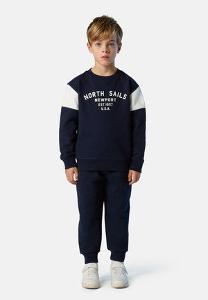 Sudadera navy con paneles de hombro blancos y texto "North Sails Newport", combinada con pantalones navy a juego y zapatillas blancas.