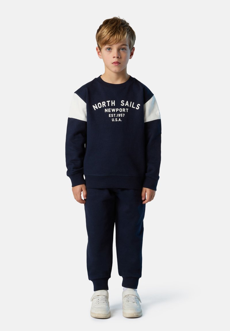 Marineblaues Sweatshirt mit weißen Schulterpanels und dem Text "North Sails Newport", kombiniert mit passenden marineblauen Hosen und weißen Sneakers.