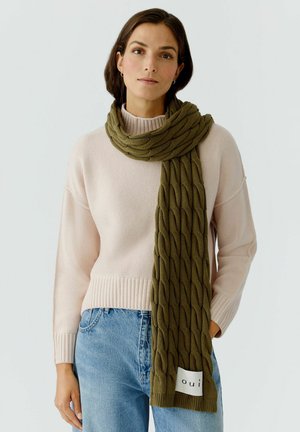 Olive green knitted scarf featuring a textured cable pattern, worn with a light pink ribbed sweater and blue denim jeans.