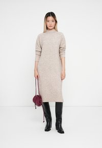 Abito midi a maglia beige con collo alto e polsini a coste, abbinato a stivali neri fino al ginocchio e a una borsa bordeaux con coulisse.