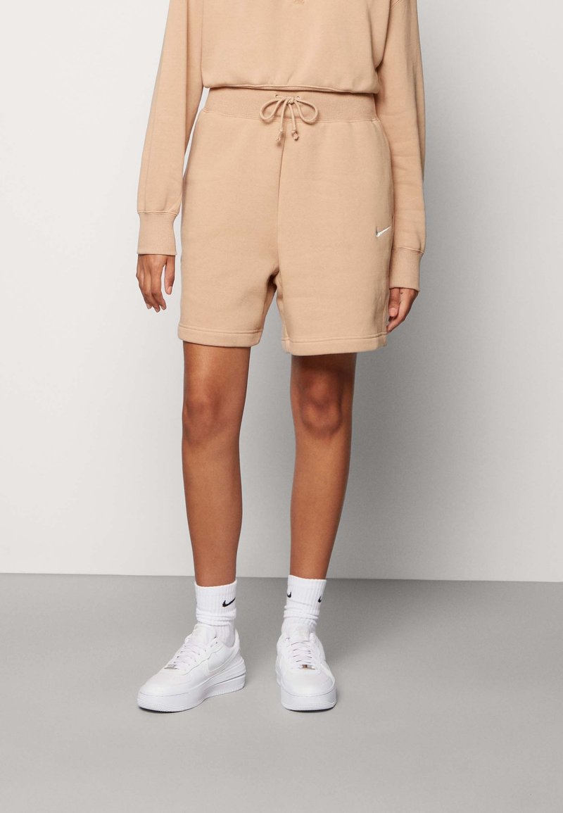Personne portant un short beige à cordon avec un petit logo latéral, un haut à manches longues assorti, des chaussettes blanches montantes et des baskets blanches, debout sur un sol gris.