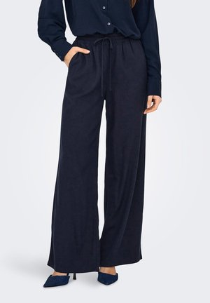 Femme portant un pantalon noir ample avec taille à cordon, chemise noire à manches longues et chaussures noires à talons, debout avec une main dans la poche.