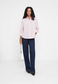 Femme avec de longs cheveux bouclés portant une chemise rose pâle boutonnée, un jean ample bleu foncé, des chaussures noires pointues et tenant un sac à main blanc.