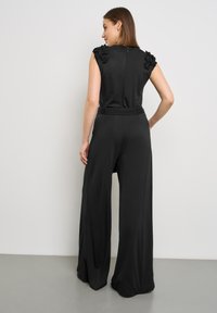 Schwarzer Jumpsuit mit weiten Hosenbeinen, floralen Applikationen an den Schultern, tailliertem Schnitt, glatter Textur und einem dezenten Reißverschluss auf der Rückseite.