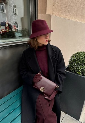 Pochette en cuir bordeaux avec quincaillerie dorée et logo, portée avec un col roulé assorti, un manteau noir et des gants, assise contre un banc vert.