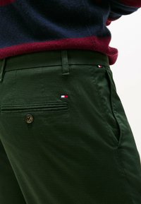 Close-up van groene textuurbroek met een knoopzak aan de achterkant en kleine rood, wit en blauw logo-tags, gecombineerd met een marineblauwe en bordeauxrode trui.