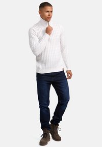 Weißer, gestrickter Pullover mit hohem Kragen und Reißverschluss, kombiniert mit dunkelblauen Jeans und braunen Schnürstiefeln.