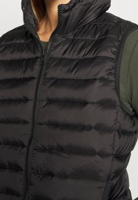 Svart vadderad puffer-väst med ståkrage, frontdragkedja och strukturerad tyg. Har sidpaneler som ger en figursydd look.