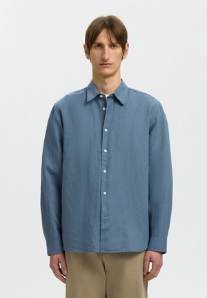 Jeune homme aux cheveux bruns lisses portant une chemise bleue à manches longues boutonnée et un pantalon beige, debout devant un fond clair uni.