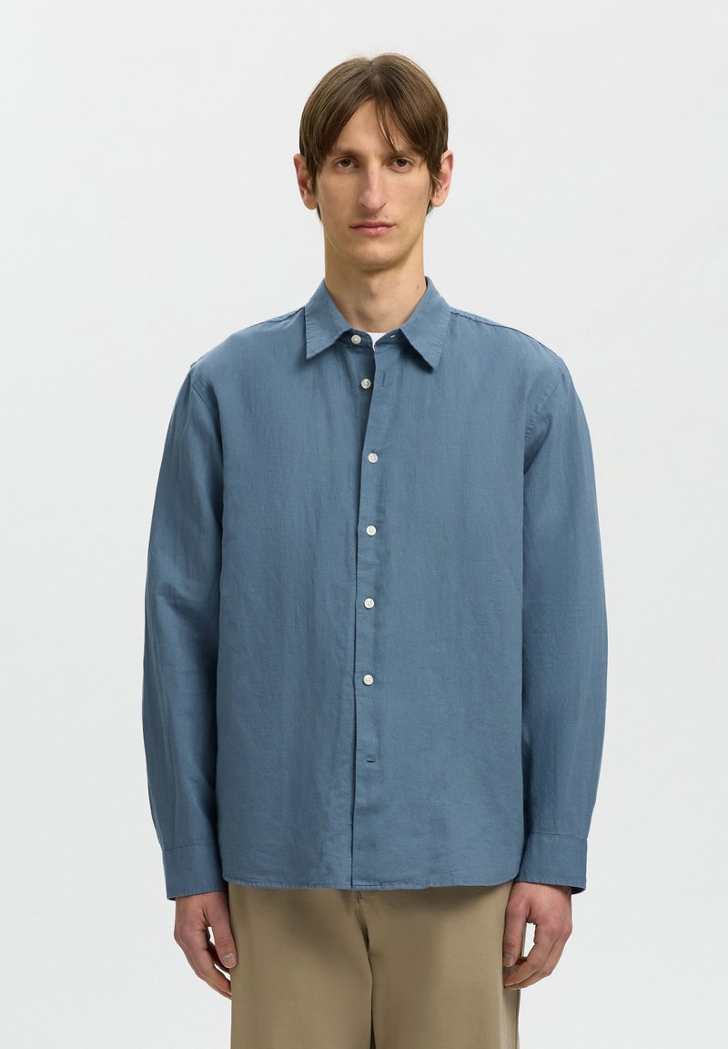 Jeune homme aux cheveux bruns lisses portant une chemise bleue à manches longues boutonnée et un pantalon beige, debout devant un fond clair uni.