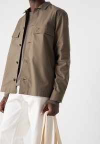 Lehká khaki bunda s knoflíkovým zapínáním vpředu, dvěma kapsami na hrudi a mírně volnějším střihem, nošená přes texturovaný svetr a bílé kalhoty.