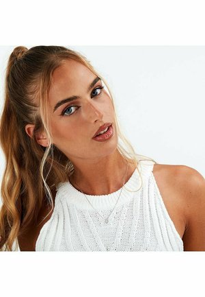 Weißes gestricktes ärmelloses Oberteil mit strukturiertem Muster, getragen mit einer zarten silbernen Halskette. Das Modell hat lange blonde Haare und blaue Augen.