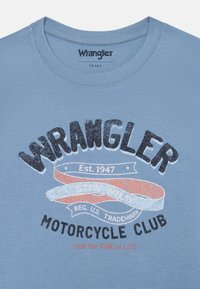 Lyseblå bomulls T-skjorte med en svart, glitrende "Wrangler"-logo, med røde og hvite detaljer og tekst: "Est. 1947," "MOTORCYKLEKLUBB," og "FOR LIVETS TUR."