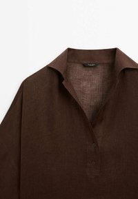 Chemise en lin marron foncé avec un col et deux boutons sur le devant, montrée de la poitrine supérieure jusqu'aux épaules sur fond blanc.