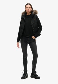 Schwarze gepolsterte Weste mit Fellkapuze, kombiniert mit einem schwarzen Strickpullover und dunklen Jeans. Das Model trägt schwarze Schnürstiefel. Minimalistischer Hintergrund.