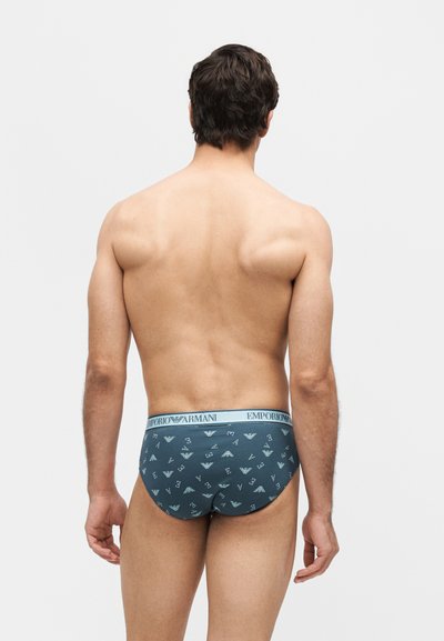 Άνδρας μοντέλο που στέκεται με την πλάτη, φοράει μπλε εσώρουχα Emporio Armani με διάσπαρτο σχέδιο λογότυπου και ανοιχτό μπλε μέση.