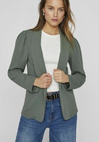 Zelený blazer se strukturovanými rameny, otevřeným předním designem, jemnou texturou, spárovaný s bílým topem a modrými džíny s vysokým pasem. Součástí je černý opasek.