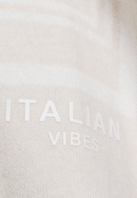 Beiger Strickstoff mit dem Text "ITALIAN VIBES" in Weiß, mit einer dezenten Textur und einem minimalistischen Design.